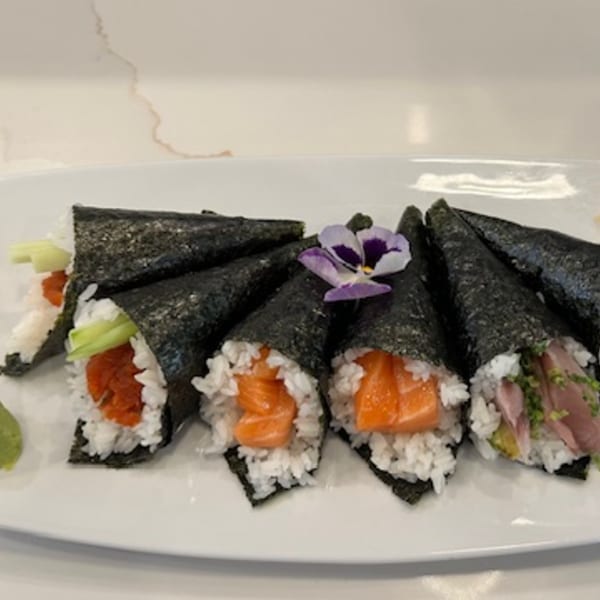 A white plate holds six sushi hand rolls, some with salmon, tuna, and avocado, garnished with a purple flower.